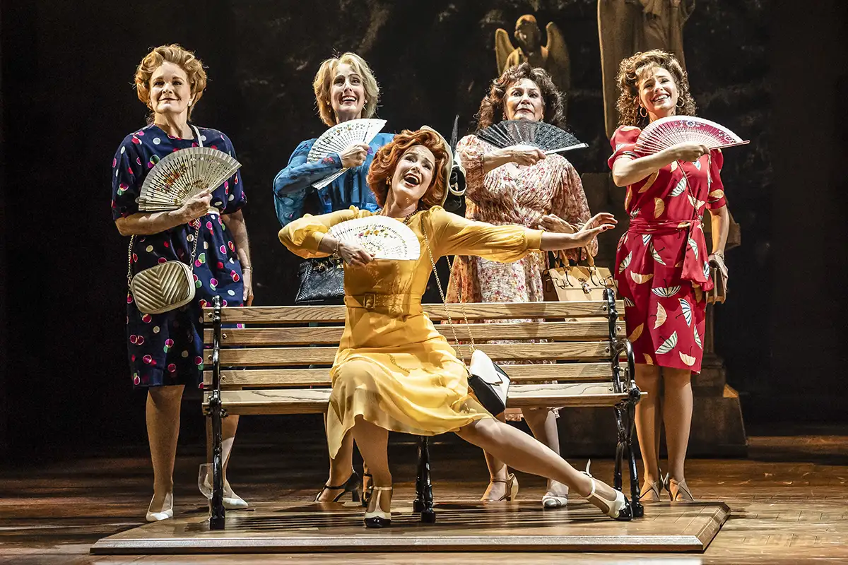 Five women in colorful vintage dresses fan themselves around a park bench