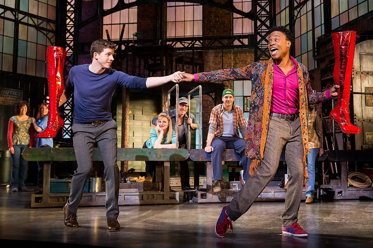 Two Kinky Boots performers dance and shake hands on the factory set while the ensemble watches, one holding red boots